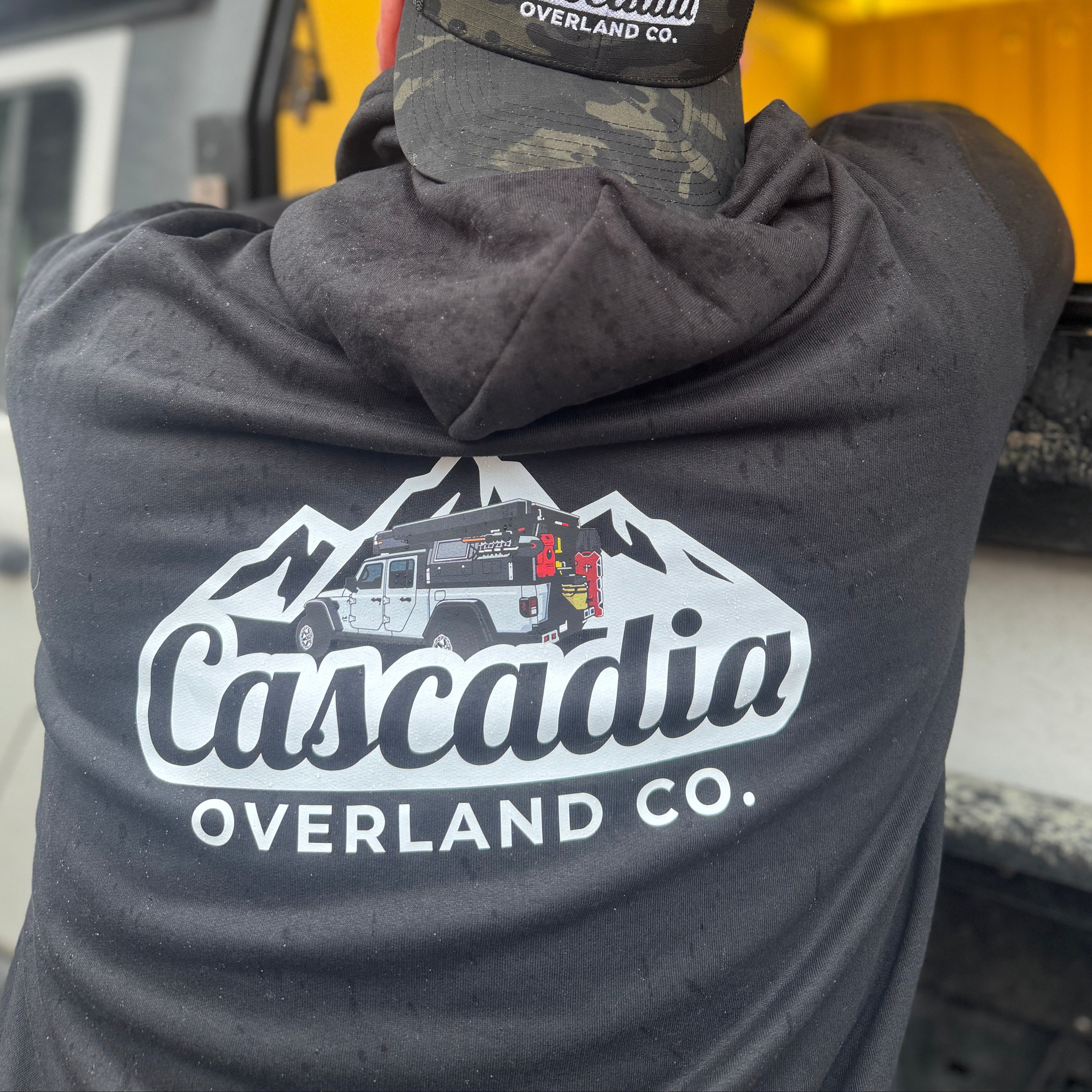 Person wearing a hoodie and cap with 'Cascadia Overland Co.' logo in front of a vehicle.