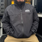 Man wearing a dark jacket with a logo, sitting in the back of a large vehicle.