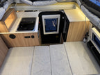 Compact kitchen area inside a vehicle with a stove, sink, and storage compartments.